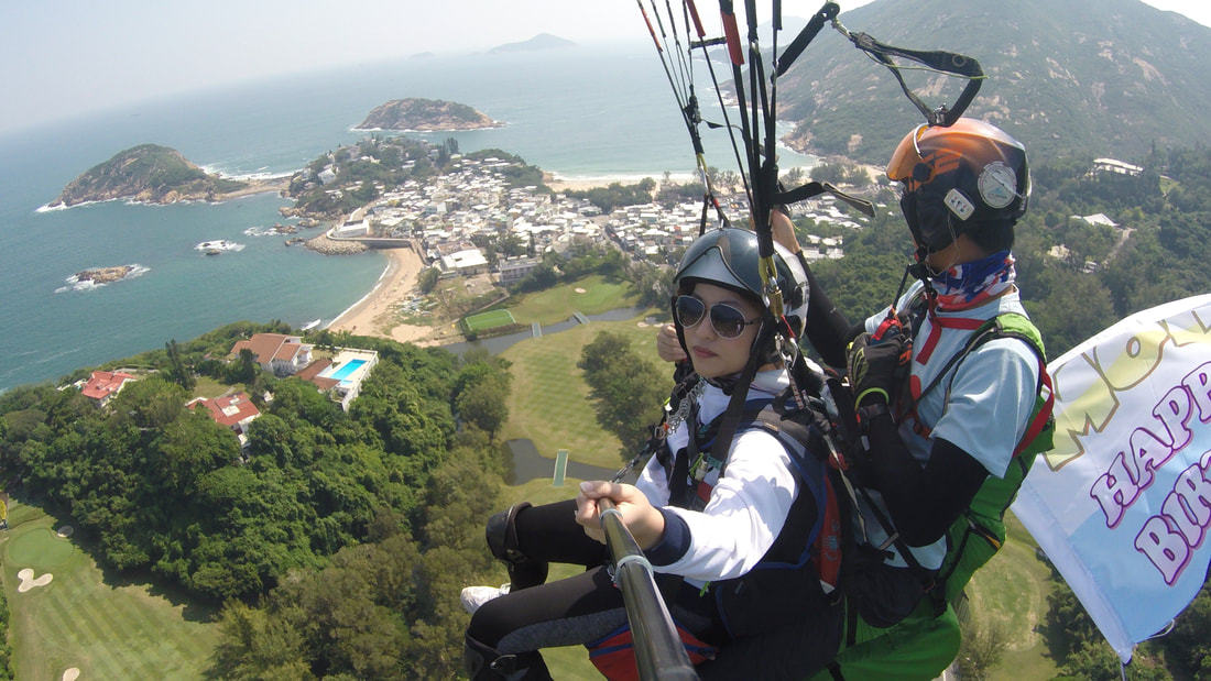 Tandem paragliding in Hong Kong over the coastline and beaches, with a passenger and pilot flying above green hills and the sea.